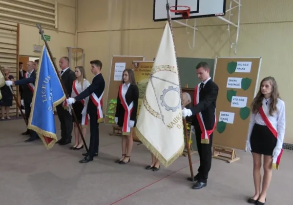 Uczniowie w galowych strojach z biało-czerwonymi wstęgami stoją w hali gimnastycznej i trzymają flagi szkolne podczas inauguracji roku szkolnego 2017/18 w Gąbinie.
