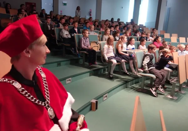 Grupa dzieci siedzi w sali wykładowej podczas inauguracji roku akademickiego Mazowieckiego Uniwersytetu Dziecięcego; na pierwszym planie osoba w czerwonym stroju ceremonialnym z dużym łańcuchem i medalem.