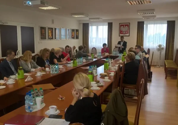 Sala konferencyjna z kilkunastoma uczestnikami szkolenia z obronności, siedzącymi przy długim stole z notatnikami, wodą i kawą, będącym w trakcie wystąpienia prowadzącego przy końcu sali.