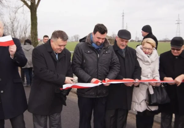 Grupa osób w zimowych ubraniach przecina czerwono‑białą wstęgę podczas uroczystości związanej z Płockim Powiatowym Uniwersytetem Trzeciego Wieku, na tle drogi i pól.