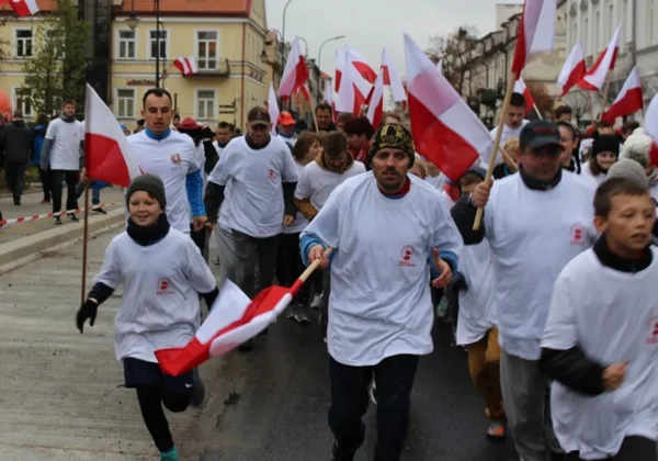 Grupa uczestników w białych koszulkach biegnie ulicą, niosąc biało-czerwone flagi Polski, na tle zabudowy miejskiej.