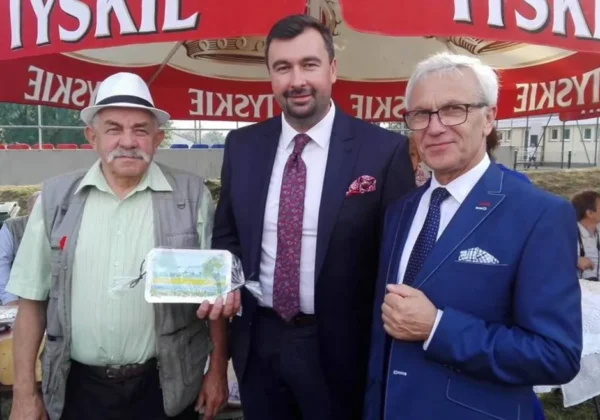 Trzech mężczyzn stoi razem na stoisku podczas Dni Małej Wsi i Dożynek Gminnych, a jeden z nich trzyma opakowany produkt.