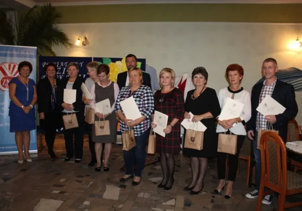 Grupa nauczycieli i pracowników samorządowych stoi na scenie podczas Powiatowego Dnia Edukacji Narodowej, trzymając dyplomy i torby z upominkami.
