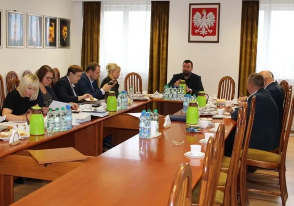 Zarząd Powiatu Płockiego podczas spotkania dotyczącego bezpieczeństwa powodziowego, siedzący przy długim drewnianym stole w sali konferencyjnej, z butelkami wody, kawą i przekąskami oraz godłem Polski na ścianie.