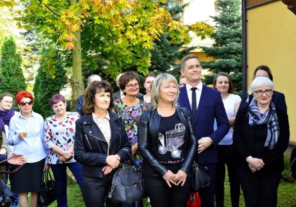 Grupa ludzi stojąca na trawniku przed żółtym budynkiem, w różnorodnych stylach ubioru, uśmiechnięta podczas wydarzenia promującego projekt „Dom z książką”.