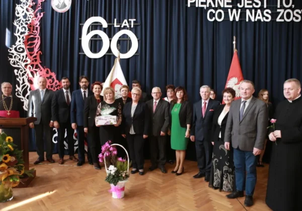 Grupa osób w formalnych strojach na scenie podczas jubileuszu 60-lecia Zespołu Szkół im. Jana Śniadeckiego w Wyszogrodzie, z flagami i kwiatami w tle.