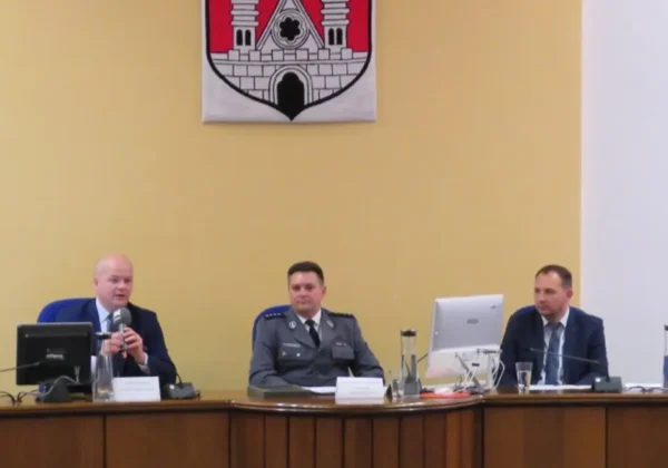 Na zdjęciu trzy osoby w garniturach i mundurze policyjnym siedzą przy długim stole podczas konferencji, a na żółtej ścianie w tle wisi herb powiatu.
