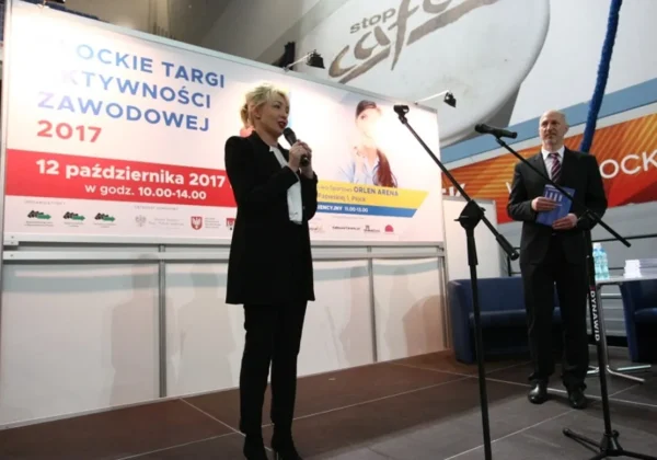 Kobieta w czarnym stroju przemawia na scenie z mikrofonem podczas Płockich Targów Aktywności Zawodowej 2017, obok niej stoi mężczyzna w garniturze, a w tle widoczny baner informacyjny wydarzenia.