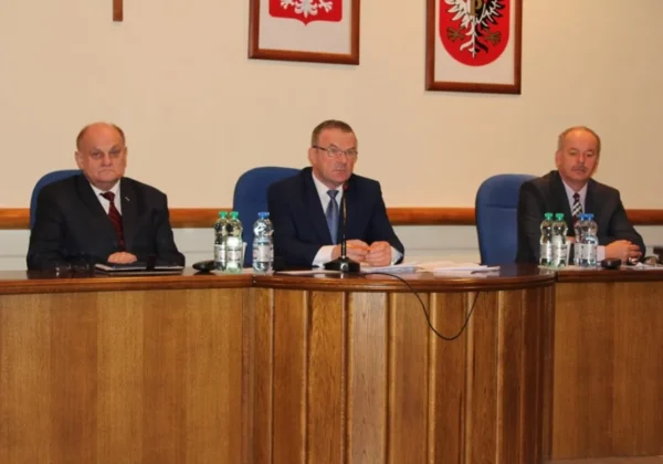 Trzech mężczyzn w garniturach siedzi za drewnianym biurkiem podczas XXVIII sesji Rady Powiatu, z mikrofonem i butelkami wody na stole, a w tle widoczne herby na ścianie.