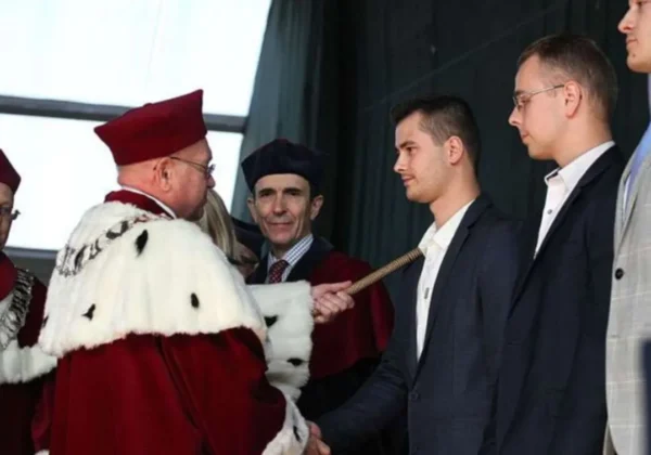 Rektor w czerwonym stroju z białym futerkiem wręcza młodemu mężczyźnie w garniturze ceremoniowy przedmiot podczas inauguracji nowego roku akademickiego w Szkole Wyższej im. Pawła Włodkowica.