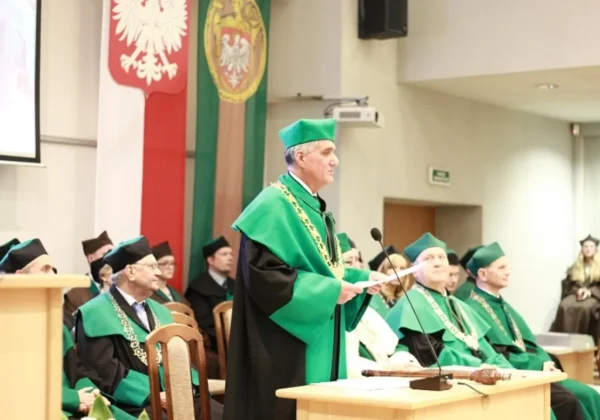 Mężczyzna w zielonej togach akademickich przemawia z mównicy podczas 51. inauguracji Politechniki Warszawskiej Filii w Płocku, otoczony innymi profesorami w zielonych togach, na tle flag i emblematów.