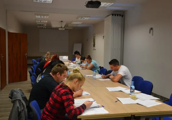 Uczestnicy i uczestniczki szkolenia umiejętności komunikacyjnych siedzą przy długim stole w sali konferencyjnej i pracują nad dokumentami w ramach projektu „Aktywizacja społeczno-zawodowa osób wykluczonych w powiecie płockim”.
