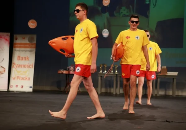 Trzech mężczyzn w żółtych koszulkach i czerwonych spodenkach, bez butów, idzie po scenie trzymając duże pomarańczowe rekwizyty podczas gali Miss i Mister Wolontariatu 2017 w Teatrze w Płocku.