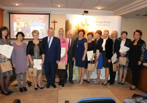 Grupa osób w eleganckich strojach stoi na sali i trzyma dyplomy oraz torby upominkowe podczas XIX Powiatowego Dnia Edukacji Narodowej.