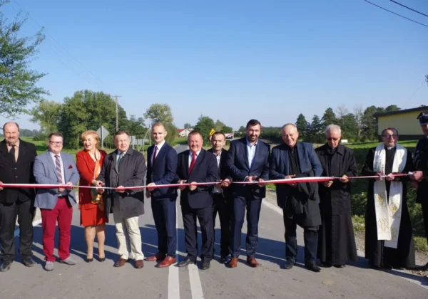 Grupa osób w formalnych ubraniach stoi na nowo wyremontowanym, blisko 3-kilometrowym odcinku drogi Białkowo-Szczytno i trzyma czerwono-białą wstęgę podczas otwarcia.