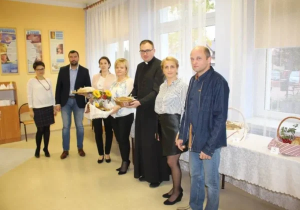 Grupa osób, w tym duchowny w sutannie, stoi w jasnym pomieszczeniu przy stole z koszami chleba i kwiatami podczas Dnia Chleba 2017.