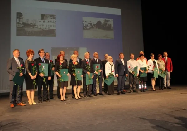 Grupa osób w formalnych ubraniach stoi na scenie podczas uroczystości z okazji 20-lecia Płockiego Pogotowia, trzymając zielone teczki i czerwone róże, z projekcją zdjęć w tle.