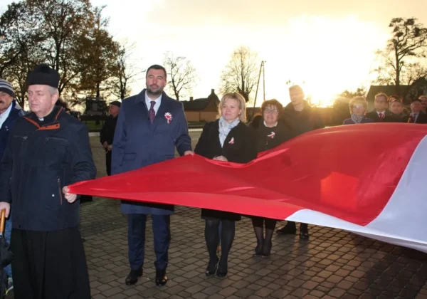 Grupa mieszkańców Wyszogrodu trzyma długi, czerwono-biały sztandar podczas Narodowego Święta Niepodległości, na tle zachodzącego słońca.