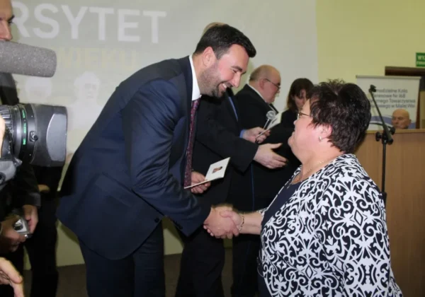 Mężczyzna w garniturze ściska dłoń starszej kobiecie podczas oficjalnej ceremonii, na tle mikrofonów i osób stojących przy scenie.