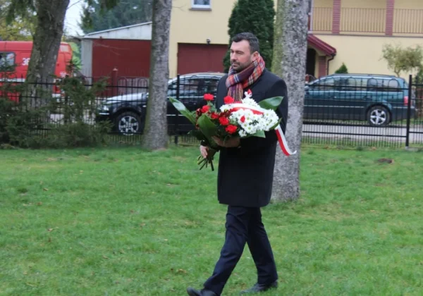 Mężczyzna w czarnym płaszczu niesie bukiet biało‑czerwonych kwiatów z czerwono‑białą wstążką podczas obchodów Święta Niepodległości w Gminie Bulkowo, na tle parku i zabudowań.