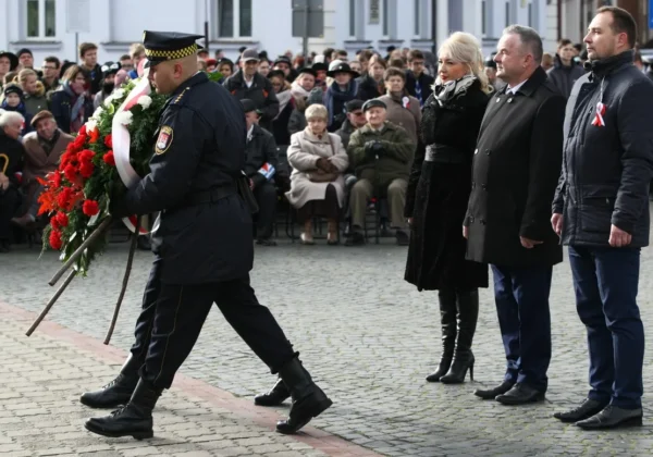 Policjant w czarnym mundurze niesie duży wieniec z czerwonych kwiatów i białą wstążką podczas Narodowego Święta Niepodległości w Płocku, przed zgromadzonymi mieszkańcami.