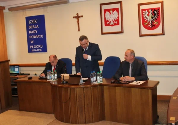 Trzech mężczyzn siedzi przy konferencyjnym stole w sali podczas XXIX Sesji Rady Powiatu w Płocku; jeden stoi i przemawia do mikrofonu, na ścianie widoczne są krzyż i dwa herby.