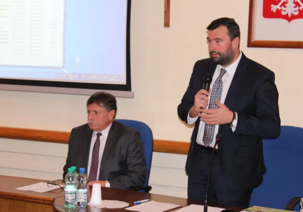 Podczas wspólnego posiedzenia Powiatowego i Miejskiego Zespołu Zarządzania Kryzysowego w Płocku stoi przy mikrofonie mężczyzna w garniturze, podczas gdy drugi mężczyzna siedzi przy stole obok.