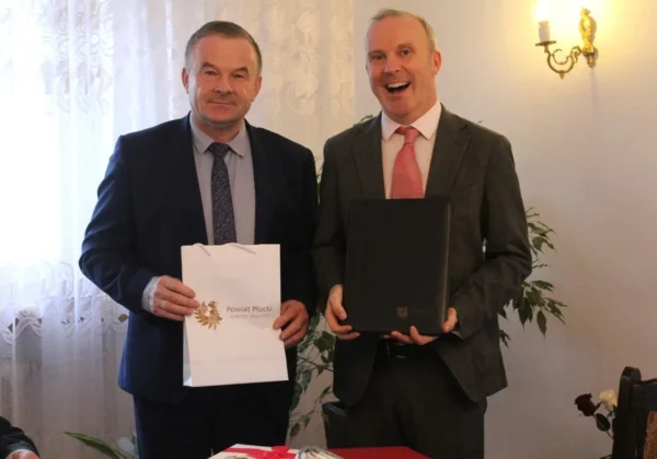 Dwóch mężczyzn w garniturach stoi obok siebie w eleganckim wnętrzu, jeden trzyma białą torbę z logo Powiatu Płockiego, drugi czarną teczkę, oboje uśmiechnięci.