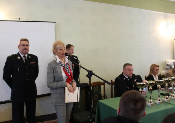 Mówczyni w szarym garniturze przemawia do mikrofonu podczas wręczania nagród IX edycji konkursu ekologicznego dla OSP i MDP, przy stole z licznymi pucharami.