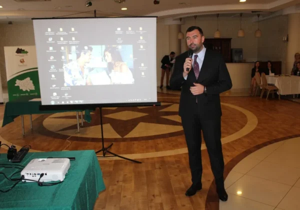 Mężczyzna w garniturze przemawia do mikrofonu podczas V Konferencji Partnerstwa Lokalnego powiatu płockiego, stojąc obok projektora i ekranu w sali konferencyjnej.