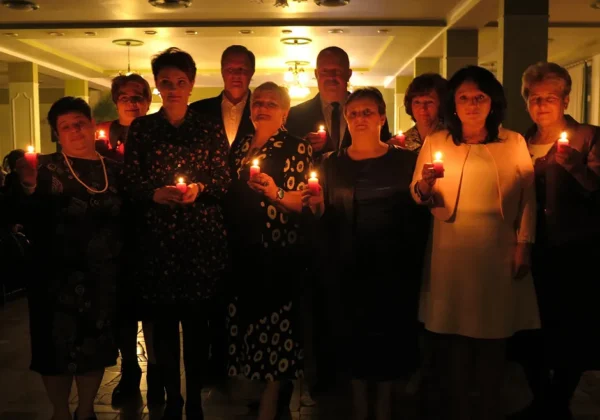 Grupa osób stojących w półmroku sali, trzymających zapalone świece podczas wigilijnego spotkania w Nowym Duninowie.