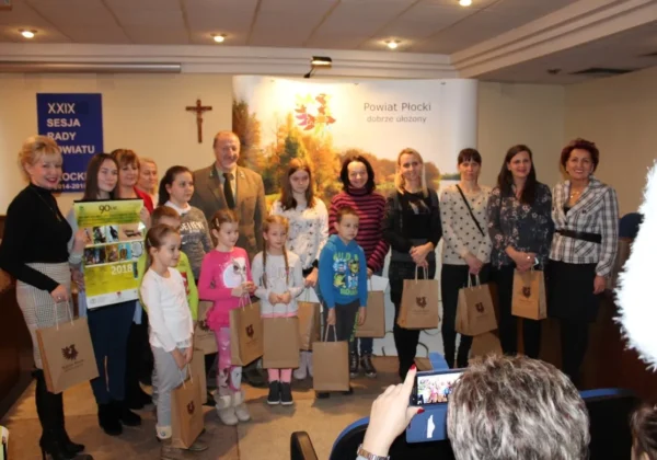 Grupa dorosłych i dzieci stoi na scenie podczas podsumowania konkursu ornitologicznego Ptaki gatunków drapieżnych, trzymając torby z logo Powiatu Płockiego.