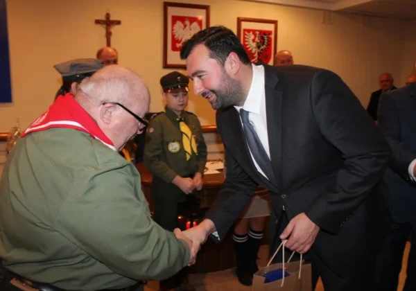 Podczas XXX sesji Rady Powiatu mężczyzna w garniturze ściska dłoń starszemu mężczyźnie w zielonej kurtce, na tle sali z polskimi herbami i osobami w mundurach harcerskich.