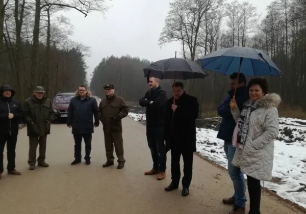 Grupa osób w zimowych kurtkach stoi na drodze w trakcie remontu w Gminie Nowy Duninów, część z parasolami, w tle lasy i resztki śniegu.
