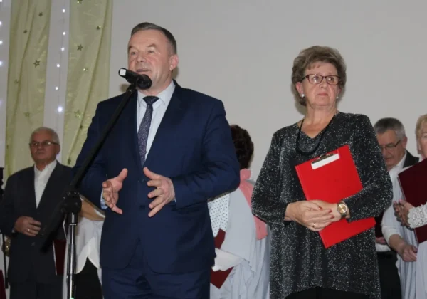 Mężczyzna w granatowym garniturze przemawia do mikrofonu na scenie, stojąc obok niego kobieta w błyszczącej bluzce trzymająca czerwony folder podczas wigilijnego spotkania Polskiego Związku Emerytów, Rencistów i Inwalidów.