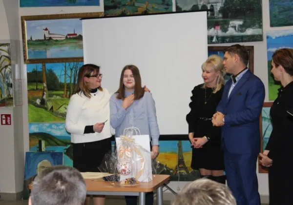 Grupa osób w sali z obrazami na ścianach i ekranem projekcyjnym wręcza upominek młodej uczestniczce podczas podsumowania projektu „Dbamy o Zdrowie – Zadbaj i TY”.