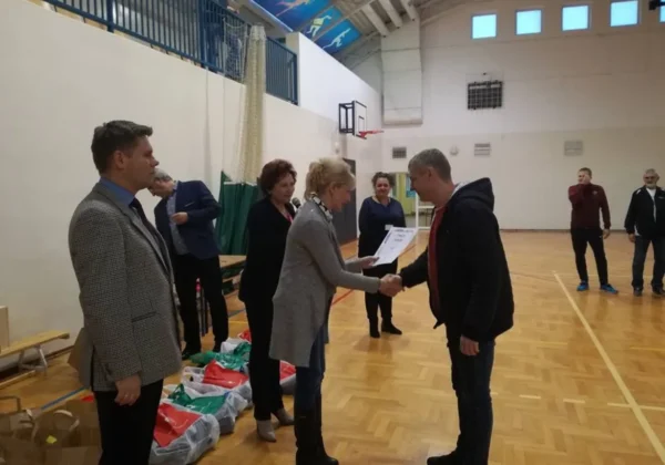 Kobieta wręcza certyfikat mężczyźnie podczas ceremonii wręczania nagród w VII Powiatowym Turnieju Piłki Siatkowej Pracowników Oświaty ZNP, na hali sportowej w towarzystwie innych uczestników.