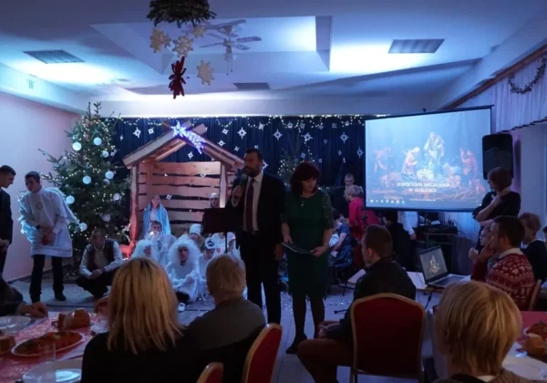 Podczas Wigilii w Zespole Szkół Specjalnych w Goślicach dwie osoby stoją przy mikrofonie i przemawiają do zgromadzonych przy stołach, w tle bożonarodzeniowa dekoracja, choinka i projekcja z napisem szkoły.