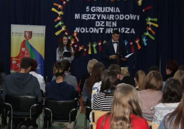 Na scenie dwójka prowadzących przemawia do publiczności podczas Międzynarodowego Dnia Wolontariusza, z banerem Powiatu Płockiego i kolorowymi dłońmi w dekoracji w tle.