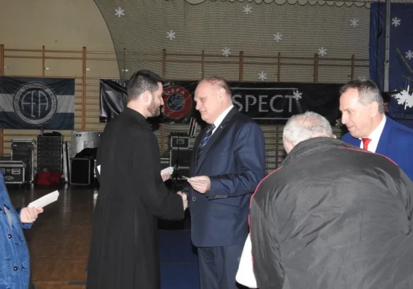 Na sali gimnastycznej podczas Wigilii gminnej w Łącku dwie osoby wręczają sobie dokumenty i ściskają dłonie, a w tle widać świąteczne dekoracje i banery.