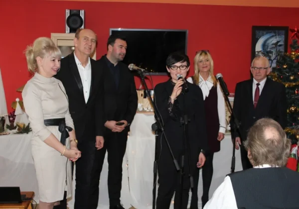 Grupa osób w formalnych strojach stoi na scenie podczas podsumowania projektu „Do szopy hej pasterze” w DPS w Koszelewie; kobieta z mikrofonem wygłasza przemówienie, obok stoją pozostali uczestnicy, a w tle widać choinkę i dekoracje świąteczne.