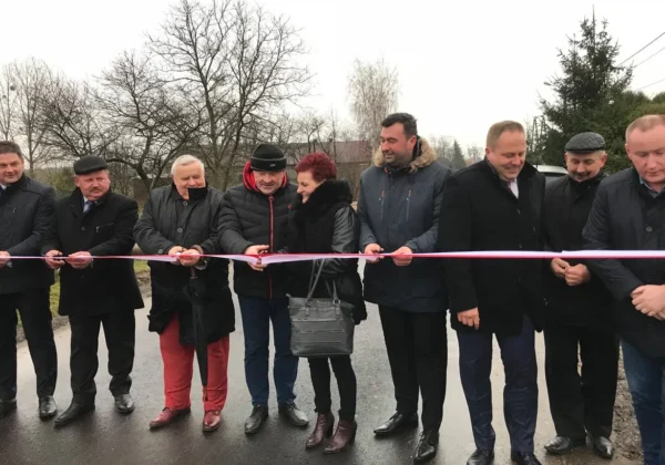 Grupa osób w zimowych kurtkach i płaszczach stoi na nowo wyremontowanej drodze powiatowej w Krubicach i przecina różową wstęgę podczas uroczystego otwarcia.