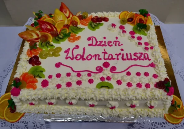 Duży biały tort ozdobiony kolorowymi owocami (kiwi, mandarynki, winogrona, pomarańcze) i kremowymi kwiatami, z napisem „Dzień Wolontariusza” różowym lukrem.