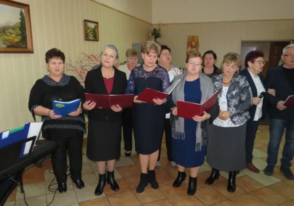 Grupa seniorów i seniorek śpiewa kolędy podczas Wigilii Senioralnej w Łęgu, trzymając czerwone śpiewniki, stojąc w sali przy pianinie po lewej.