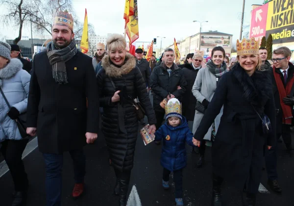 Uczestnicy Orszaku Trzech Króli idą ulicą w zimowej scenerii, noszą królewskie korony i trzymają się za ręce, prowadząc małe dziecko w błękitnej kurtce.