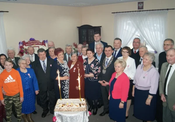 Grupa mieszkańców gminy Brudzeń Duży w eleganckich strojach stoi wokół tortu z iskrami, wręczając medale i składając gratulacje z okazji jubileuszu małżeńskiego.