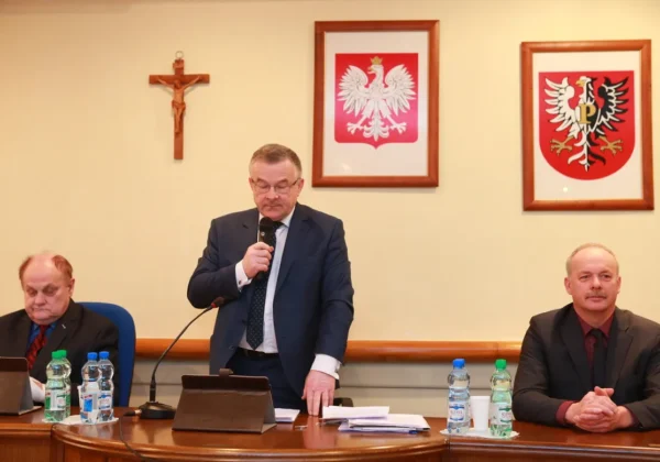 Trzech mężczyzn przy stole konferencyjnym podczas XXXI sesji Rady Powiatu; jeden stoi i przemawia do mikrofonu, w tle widoczny krzyż i dwa herby na ścianie.