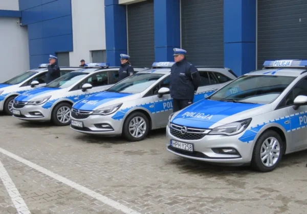 Nowe radiowozy policji w kolorze niebiesko-srebrnym stoją w rzędzie na parkingu przed budynkiem, obok których stoją funkcjonariusze w mundurach.