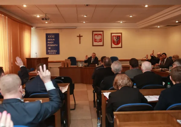 Sala obrad Rady Powiatu w Płocku podczas XXXII sesji, radni podnoszą ręce do głosowania, a na ścianie wiszą tablica z napisem sesji oraz herby.
