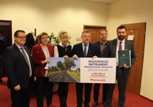 Grupa sześciu urzędników w garniturach stoi w wnętrzu i prezentuje duży plakat promujący Mazowiecki Instrument Wspierania Dróg Lokalnych.